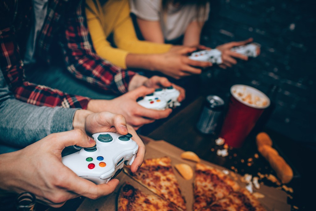 Four young people playing on gaming console, you can only see their hands. In front of them is a variety of food, including pizza, chips, and energy drinks